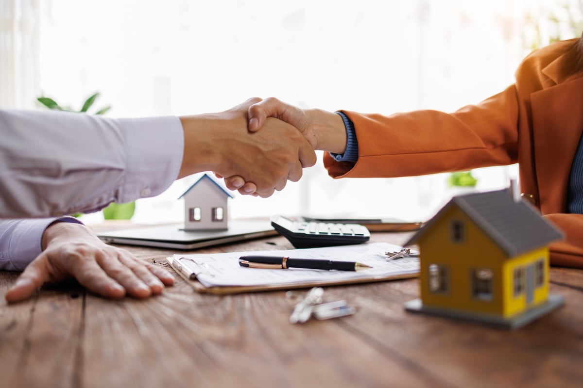 Businesspeople shaking hands over property contract, miniature houses on wooden desk